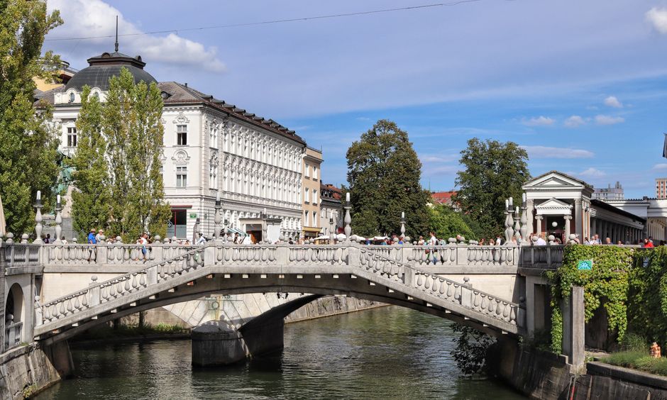 Sunny day at Ljubljana's iconic Triple Bridge with bustling pedestrians.