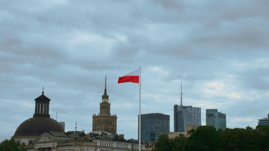Skyline of Warsaw featuring the Polish flag and iconic buildings under a cloudy sky.